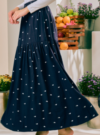 A navy blue tiered maxi skirt with a small white leaf pattern, worn by a model in a styled indoor setting with potted plants and fruit crates in the background.