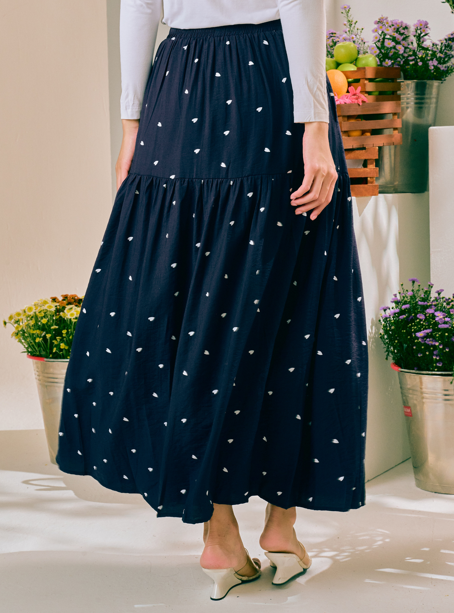 A navy blue tiered maxi skirt with a small white heart pattern, worn by a model in a brightly lit indoor setting with potted plants.
