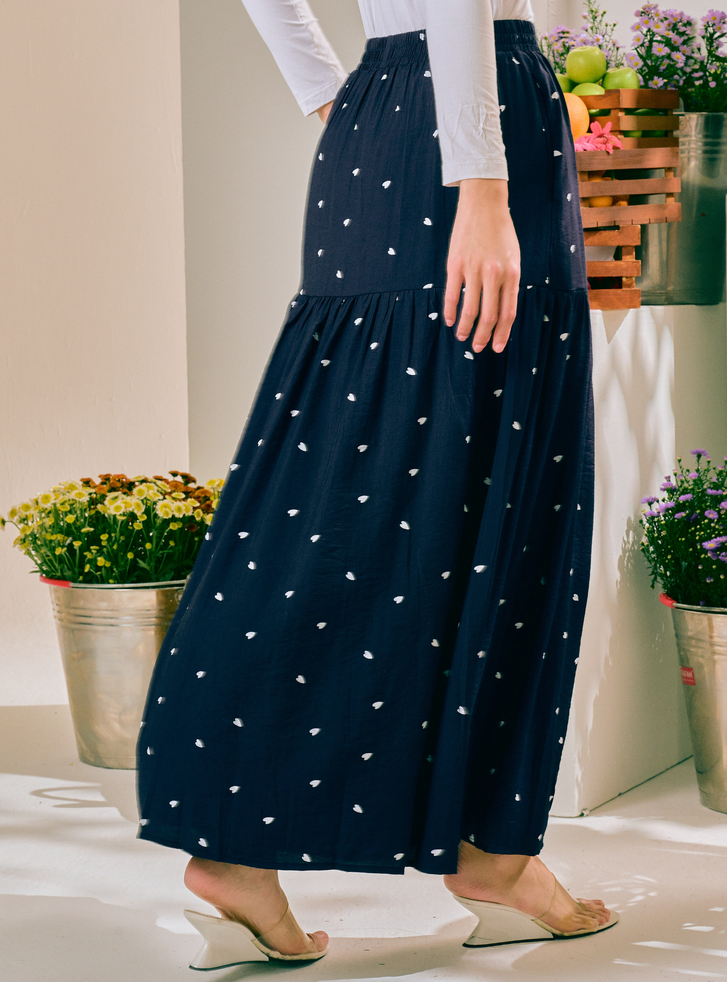 A navy blue tiered maxi skirt with a small white heart pattern, worn by a model in a brightly lit indoor setting with potted plants.