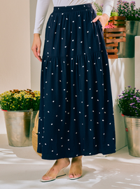 A navy blue tiered maxi skirt with small white embroidered floral motifs, worn by a model in a brightly lit indoor setting with potted plants.