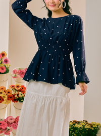 A woman wearing a navy blue long-sleeve blouse with a white ditsy floral print and a smocked waist, paired with a white tiered skirt. She is standing in a brightly lit indoor setting with potted flowers in the background.