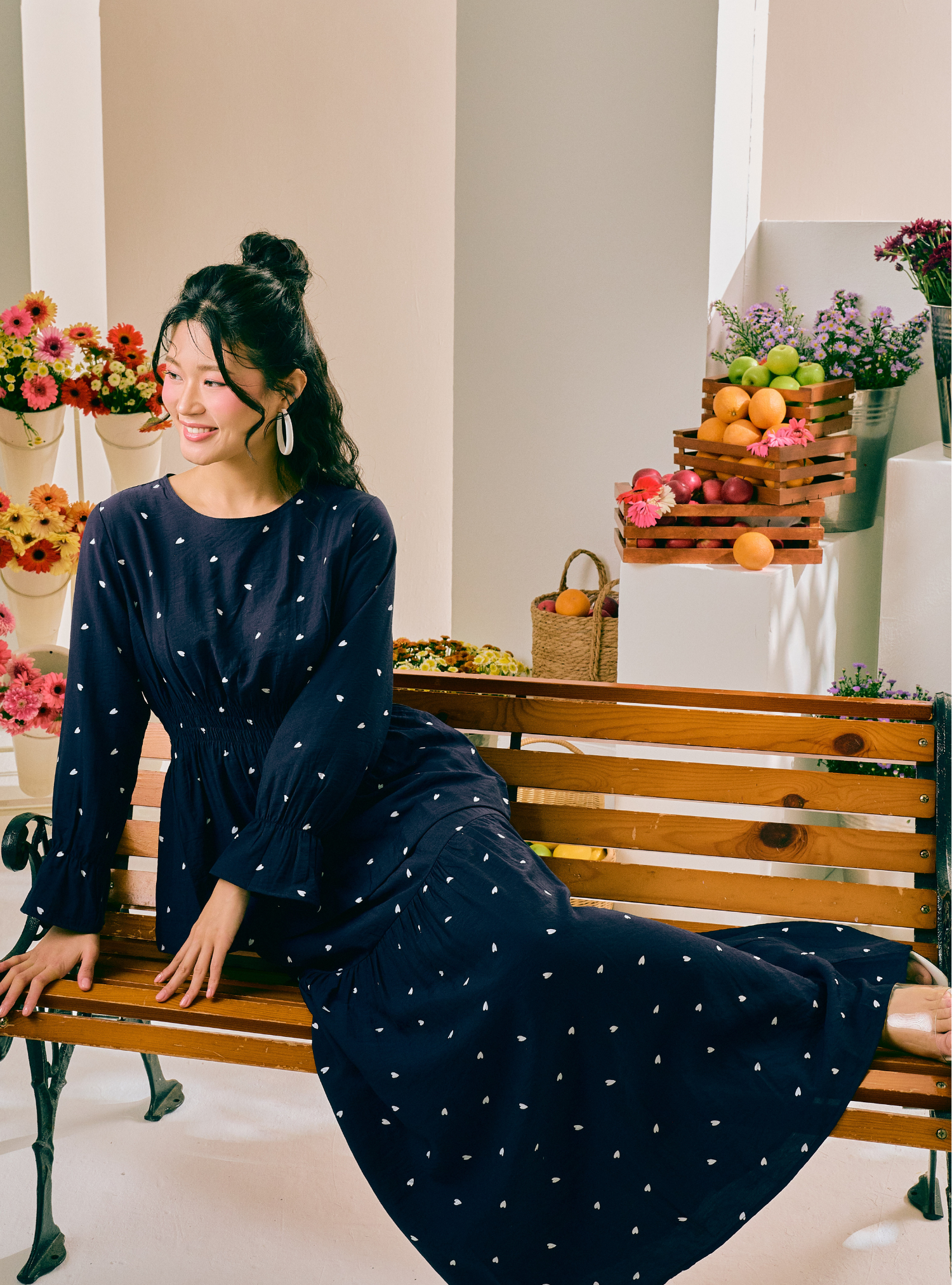 A woman wearing a navy blue long-sleeve maxi dress with small white polka dots sits on a wooden bench in a bright, styled indoor setting with flowers and fruit baskets in the background.
