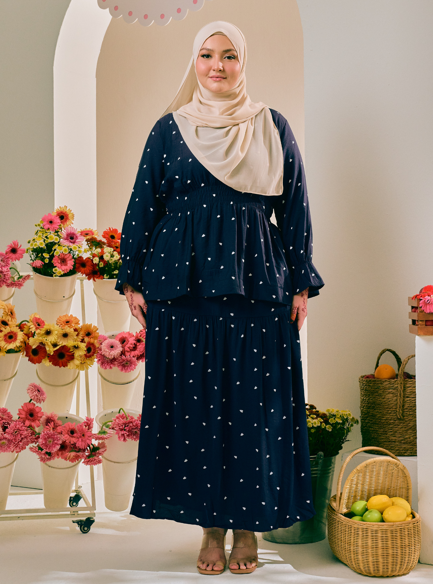 A woman wearing a navy blue two-piece modest outfit with white polka dots, featuring a long-sleeve peplum-style top and matching maxi skirt, paired with a light beige hijab. She is standing in a styled indoor setting with decorative flower arrangements and a basket of fruit.