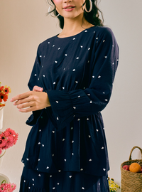 A woman wearing a navy blue long-sleeve tunic dress with a small white heart pattern, styled with large hoop earrings in a warm, neutral-toned setting with flowers and a basket.