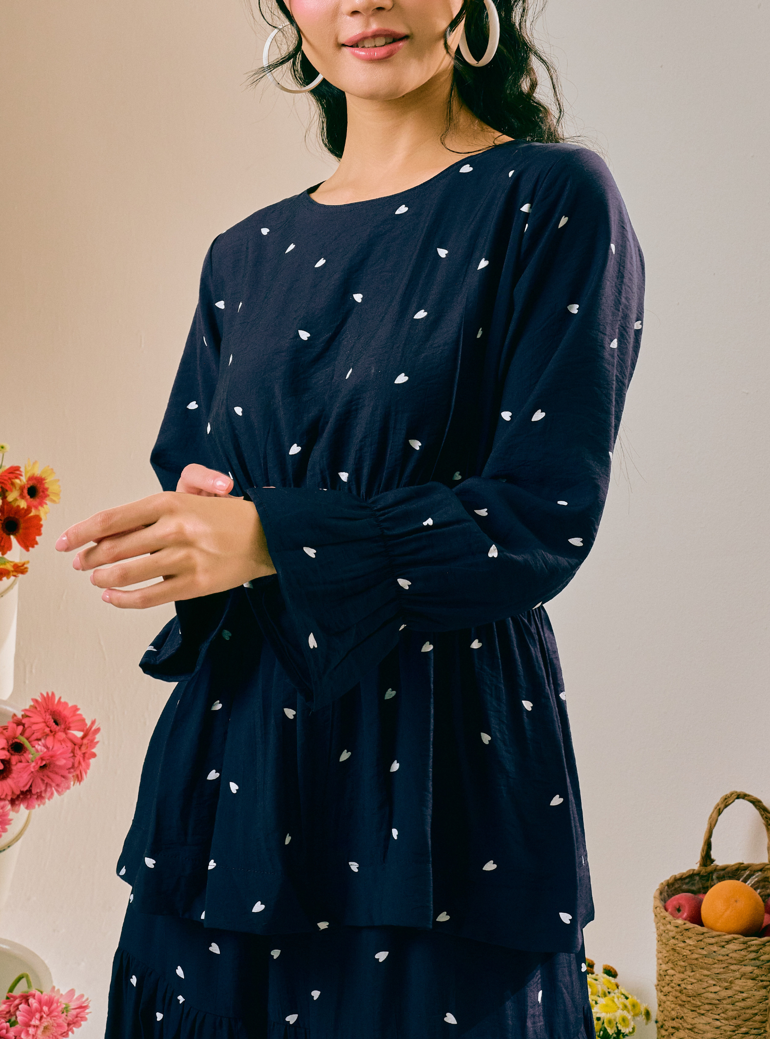 A woman wearing a navy blue long-sleeve tunic dress with a small white heart pattern, styled with large hoop earrings in a warm, neutral-toned setting with flowers and a basket.