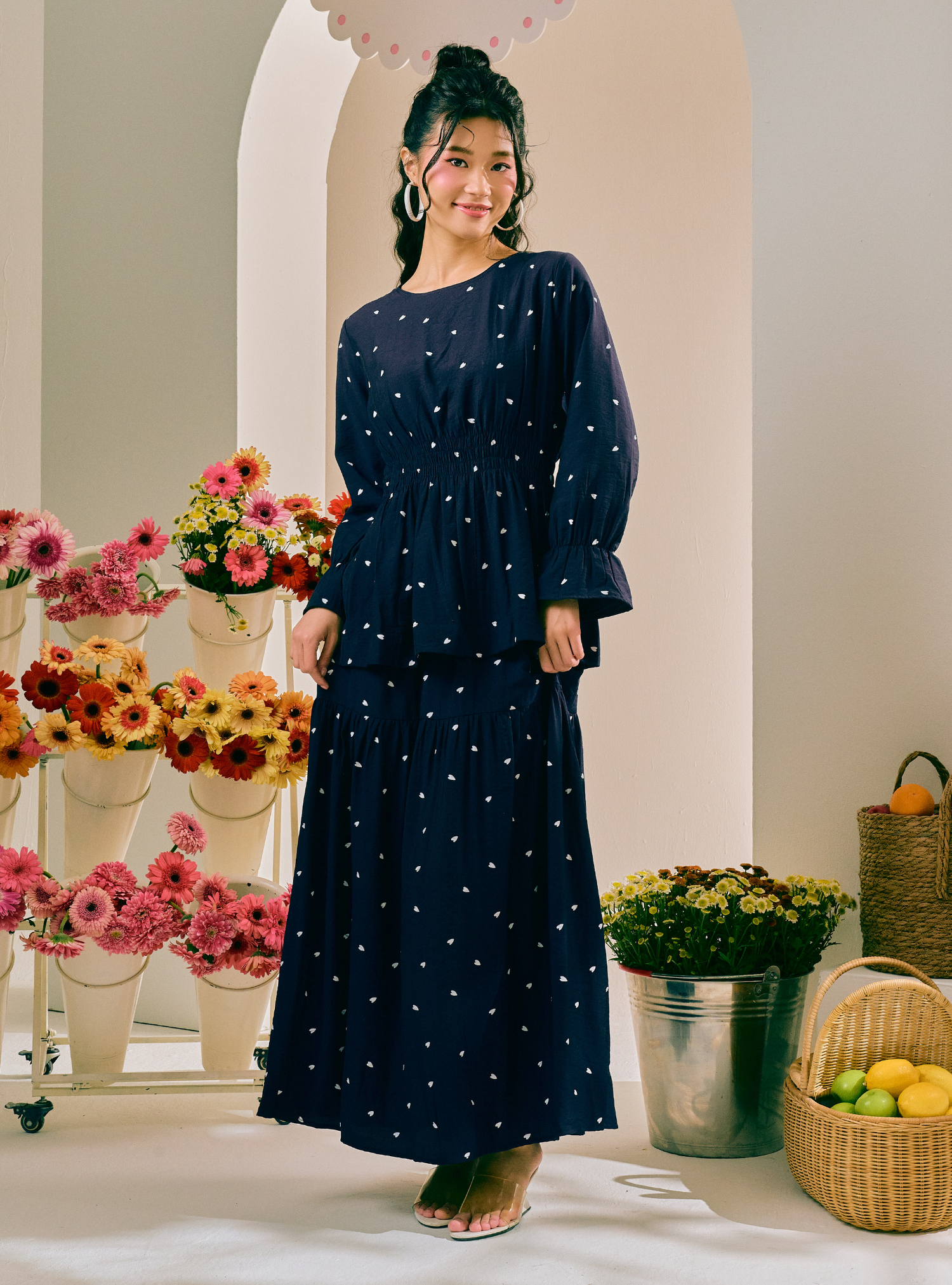 A woman models a navy blue maxi dress with white polka dots, standing in a styled indoor setting with flower arrangements and baskets.