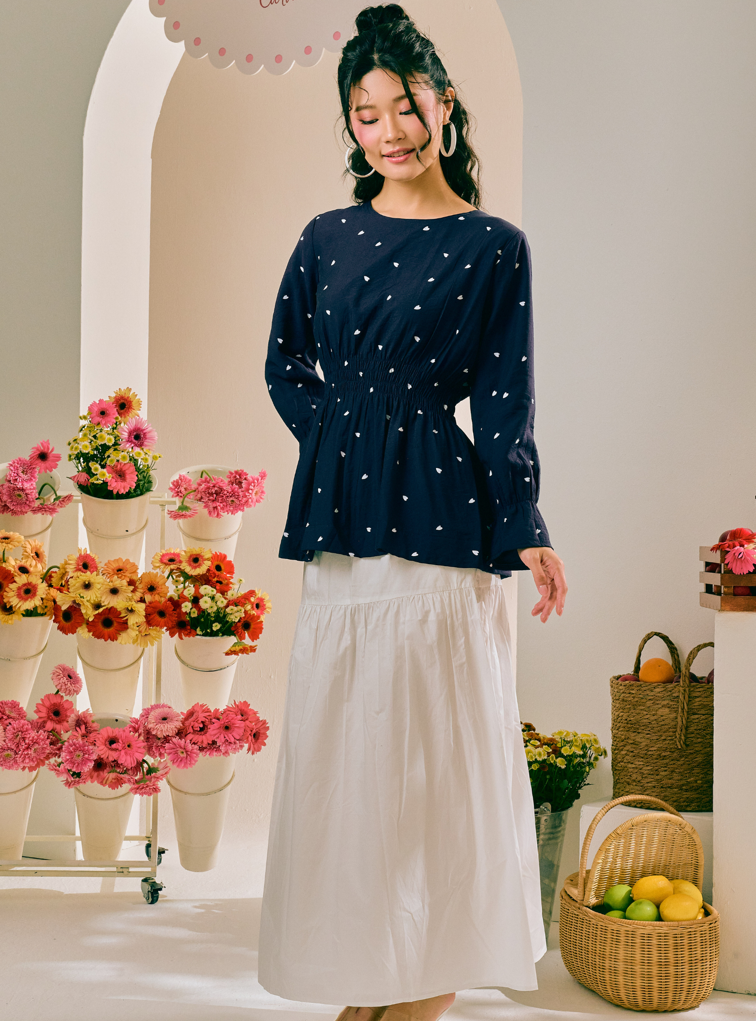 A woman stands in a bright indoor studio setting wearing a navy long-sleeve blouse with small white polka dots and a white midi skirt. The background features arches, potted flowers, and woven baskets.