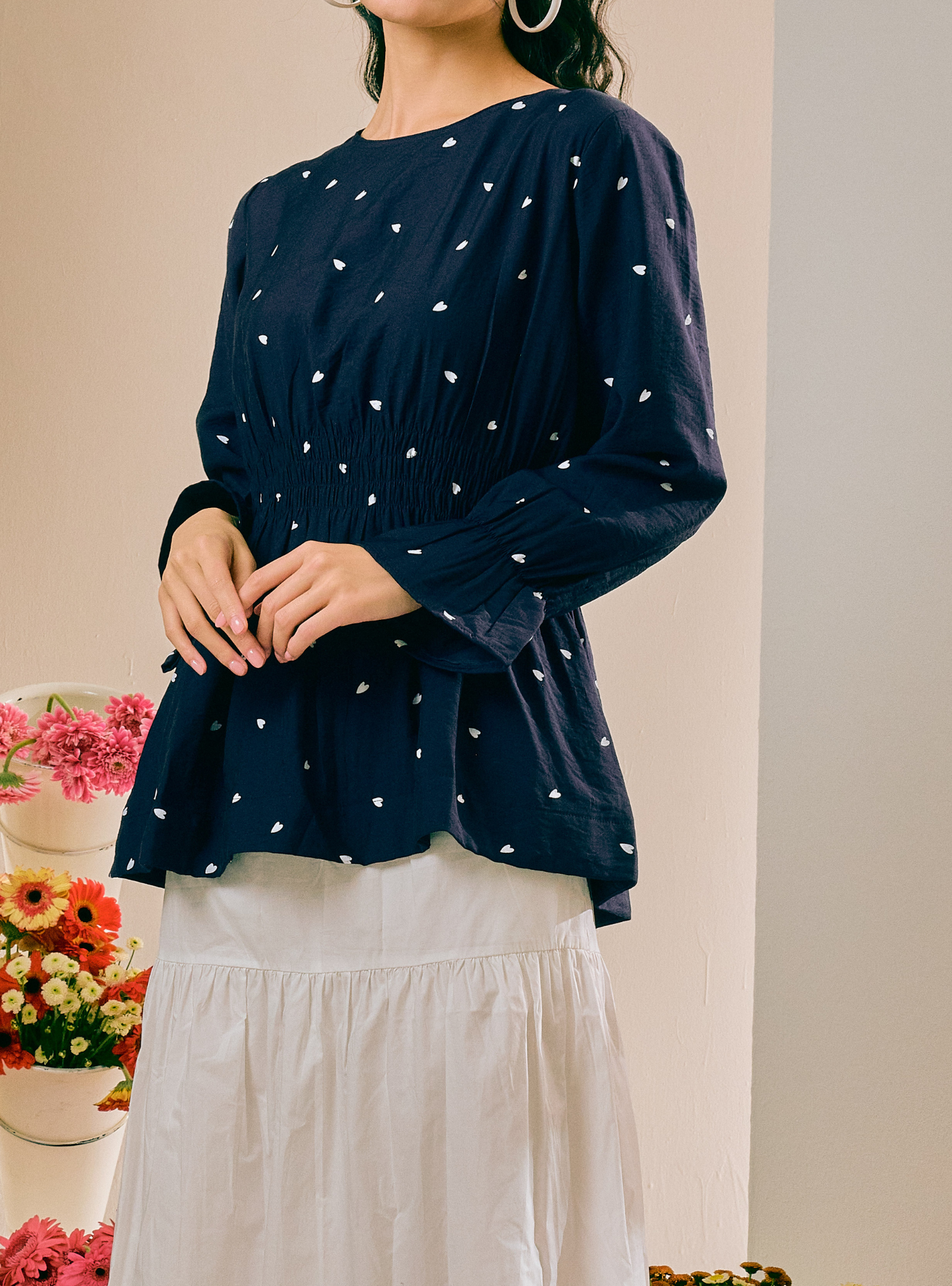 A woman wearing a navy blue long-sleeve blouse with a white heart pattern and a gathered peplum hem, paired with a white skirt. The photo is cropped from the chin down, set against a warm beige wall with decorative flowers on the left.
