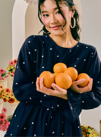 A woman wearing a navy blue long-sleeve dress with small white heart prints holds a pile of oranges in her hands. She is wearing large gold hoop earrings and standing in front of a cream-colored arched doorway with floral decorations on the sides.