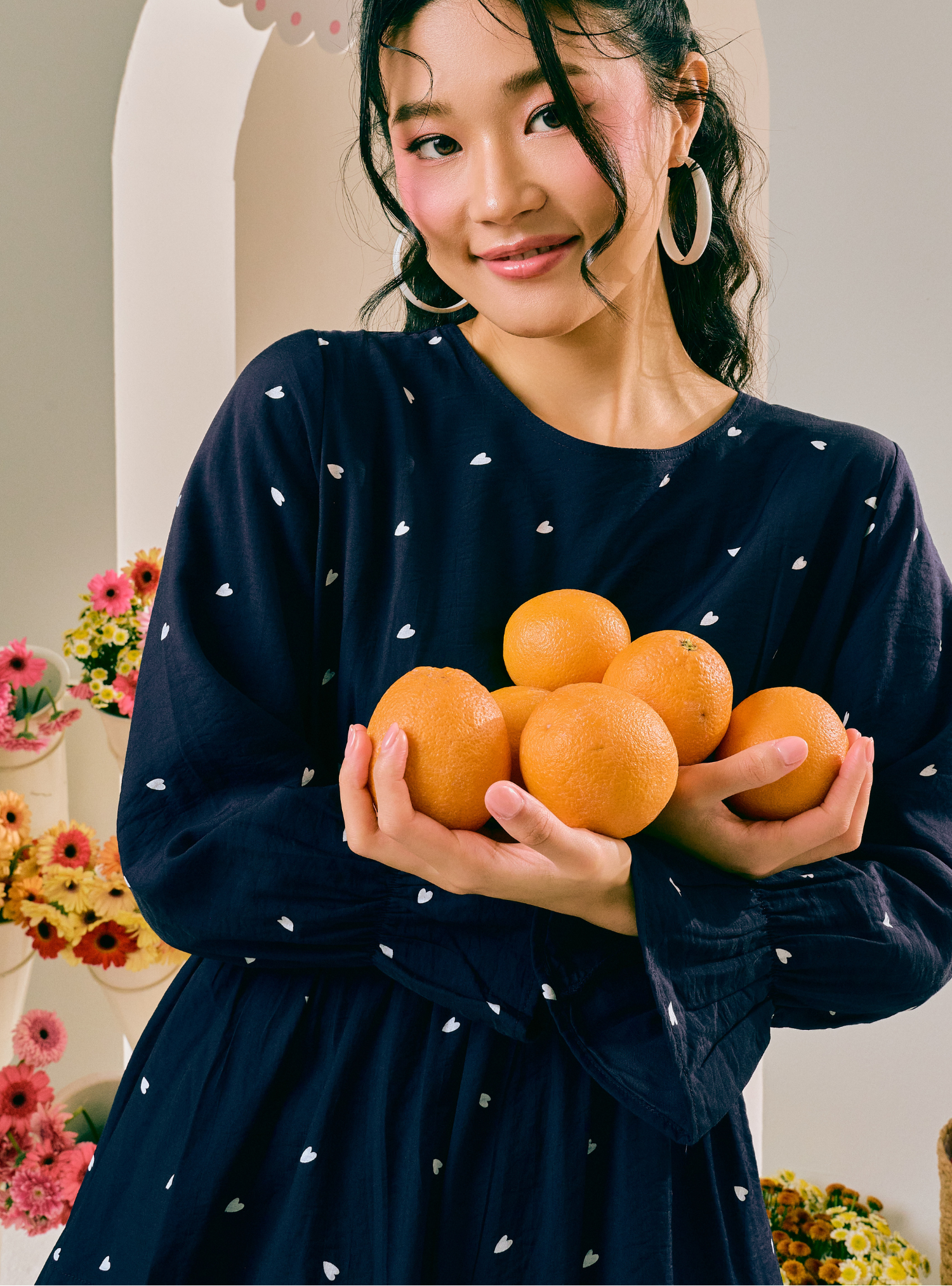 A woman wearing a navy blue long-sleeve dress with small white heart prints holds a pile of oranges in her hands. She is wearing large gold hoop earrings and standing in front of a cream-colored arched doorway with floral decorations on the sides.