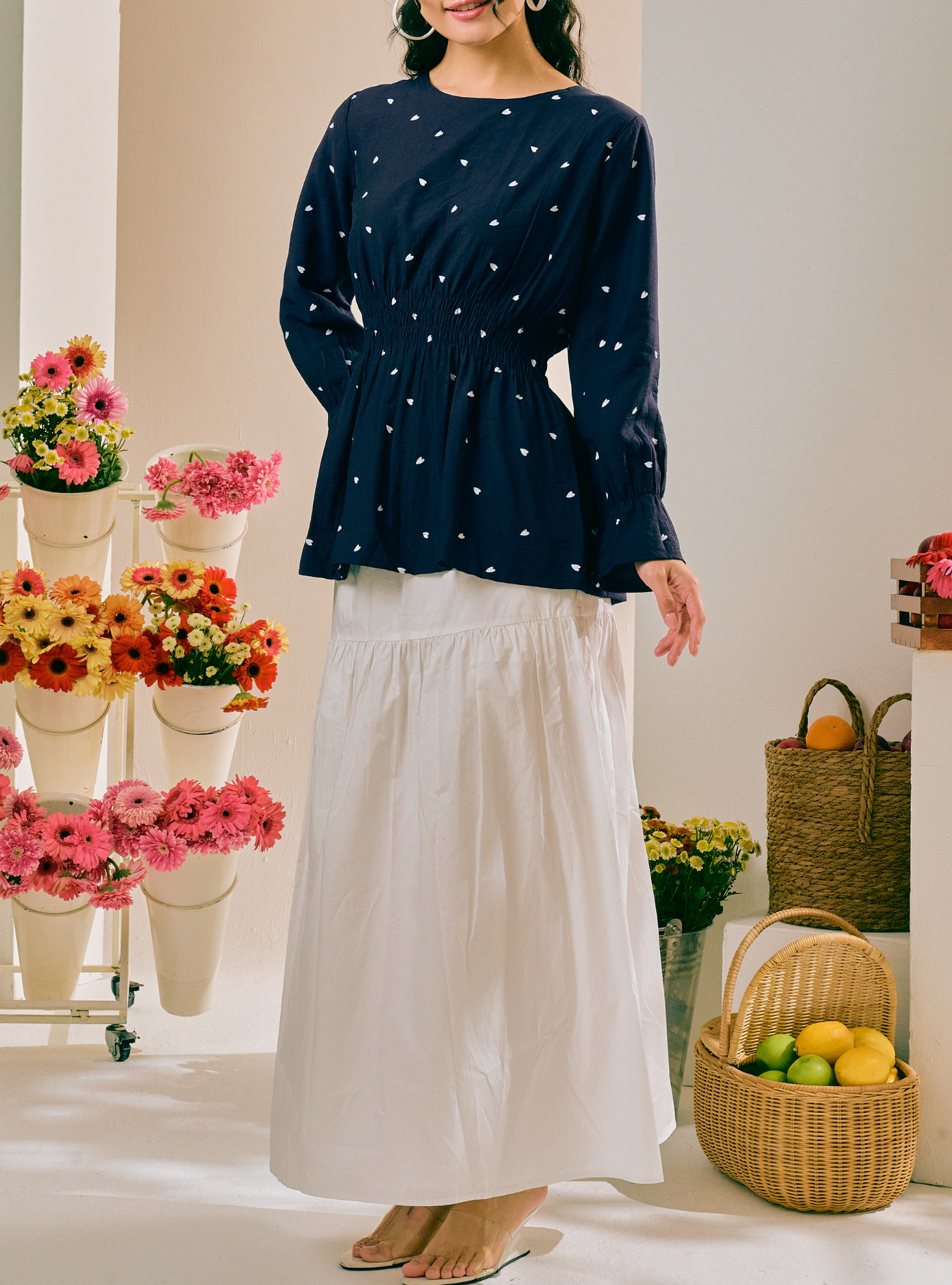 A woman models a navy blue long-sleeve peplum top with small white embroidered dots, paired with a white maxi skirt, in a bright, styled indoor setting with flowers and a wicker basket.