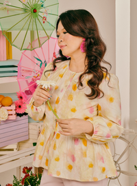A woman wearing a pale yellow long-sleeve blouse with pink and yellow floral patterns, holding a white flower. She is standing in a decorated indoor setting with a pink folding umbrella, books, and flowers in the background.