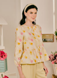 A woman wearing a cream-colored long-sleeve blouse with yellow and pink watercolor-style polka dots, paired with light yellow pants and a beige headband, standing in a brightly lit indoor setting.