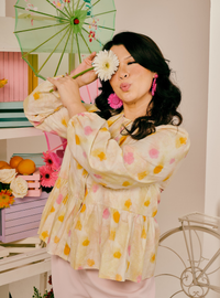 A woman posing in a cream-colored tiered blouse with yellow and pink floral patterns, holding a white daisy in front of her eye. She is styled with a green parasol and pink earrings in a brightly lit, pastel-colored room.
