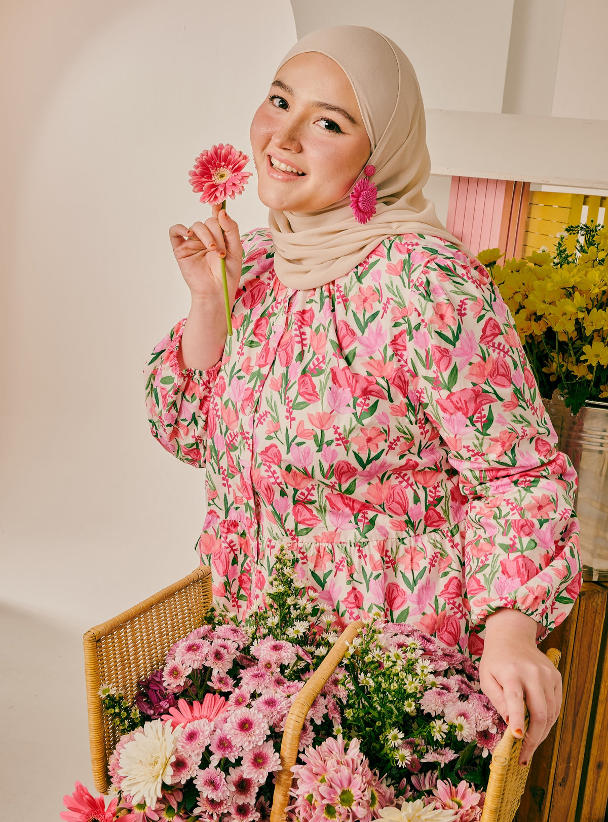 A woman wearing a beige hijab and a pink floral long-sleeve top holds a pink flower while sitting behind a wicker basket filled with pink and white chrysanthemums in a bright indoor setting.