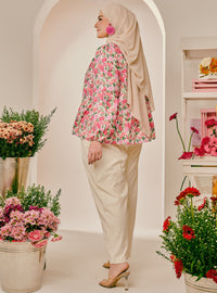 A woman wearing a beige hijab, a pink and green floral long-sleeve blouse, and cream-colored trousers stands in profile in a bright studio decorated with potted flowers.