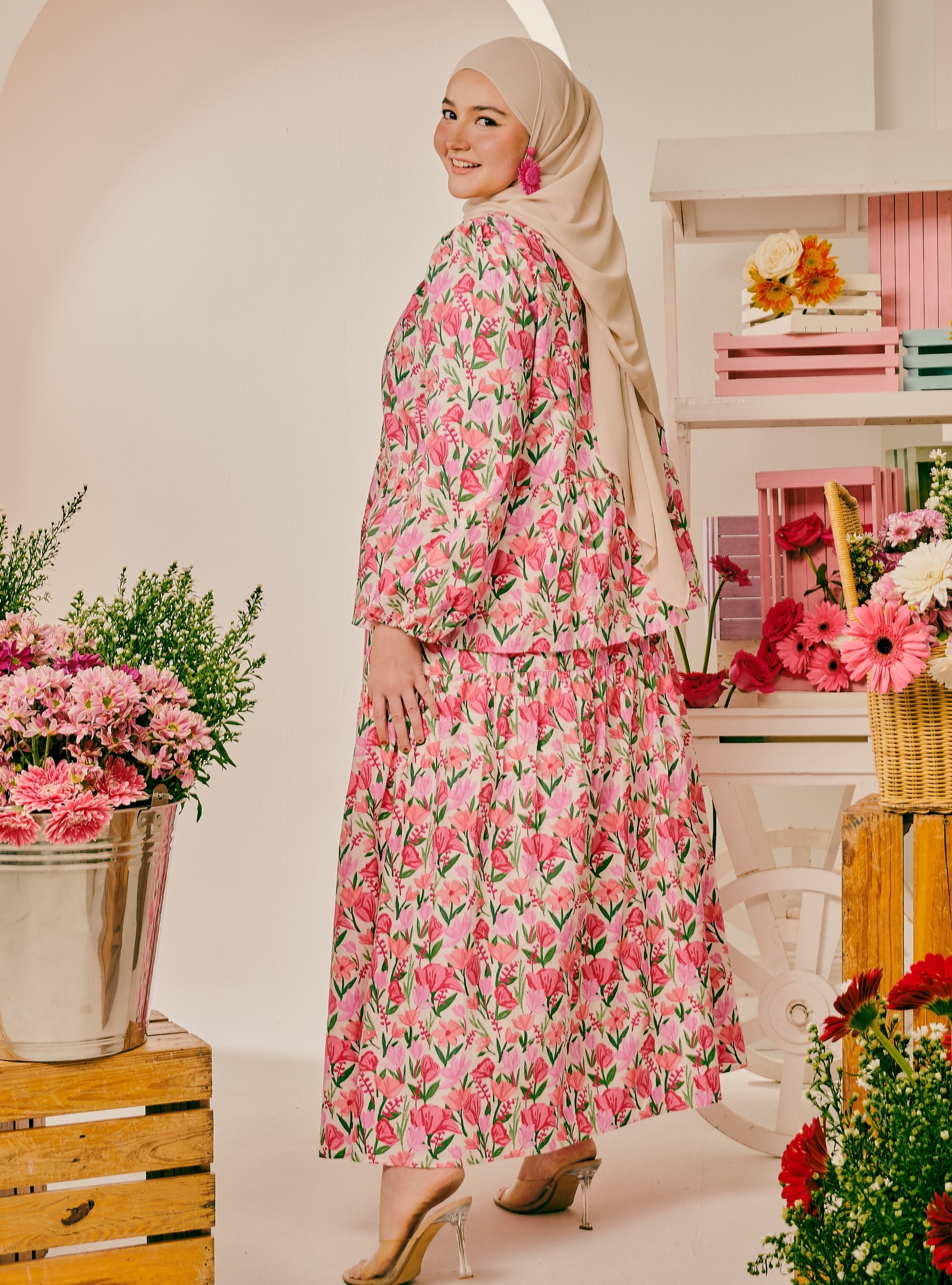 A woman wearing a pink floral long-sleeve maxi dress and a beige hijab, standing in a decorated indoor studio setting with flowers and shelves in the background.