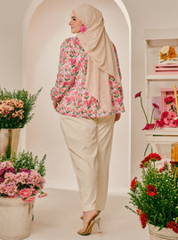 A woman wearing a beige hijab, a pink and green floral long-sleeve blouse, and cream-colored trousers, standing in a studio with arches and potted flowers.