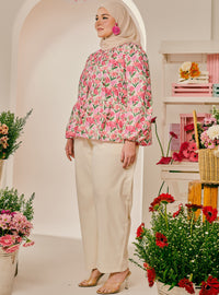 A woman wearing a pink floral long-sleeve blouse with cream wide-leg pants and a beige hijab, standing in a bright, flower-filled studio setting.