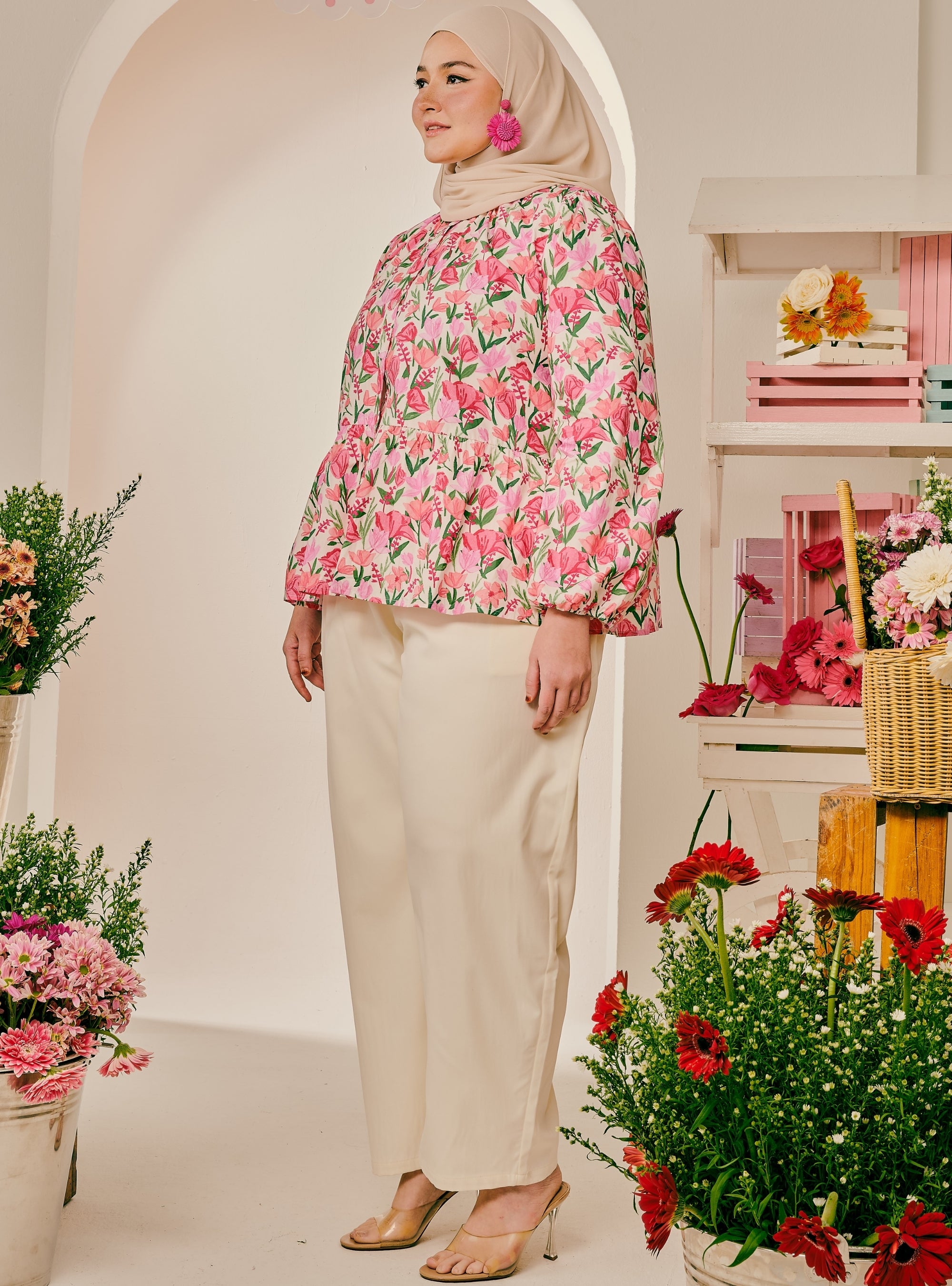 A woman wearing a pink floral long-sleeve blouse with cream wide-leg pants and a beige hijab, standing in a bright, flower-filled studio setting.