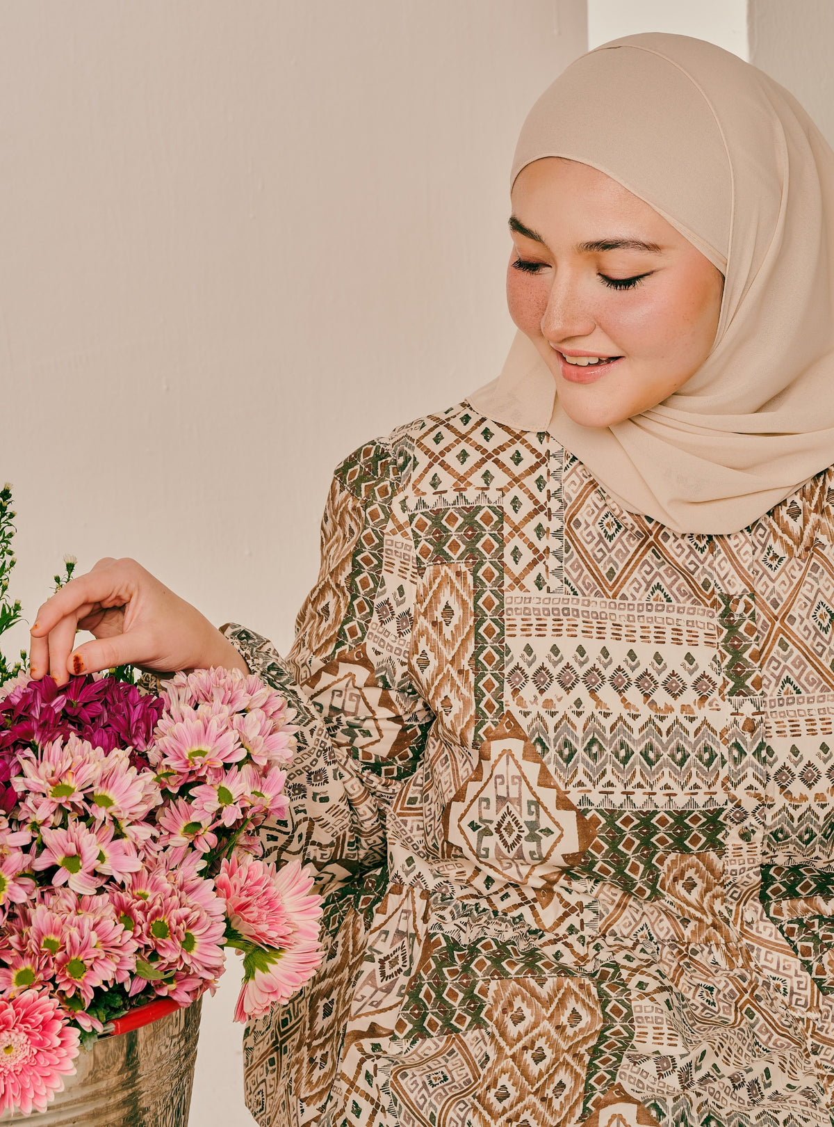 A woman wearing a beige hijab and a long-sleeved patterned blouse with geometric brown, beige, and green motifs, holding a vibrant pink flower bouquet against a neutral beige background.