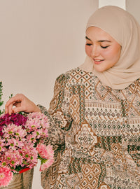 A woman wearing a beige hijab and a long-sleeved patterned blouse with geometric brown, beige, and green motifs, holding a vibrant pink flower bouquet against a neutral beige background.