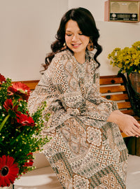 A woman wearing a long-sleeved kaftan dress with a brown, beige, and gray geometric pattern, sitting on a wooden bench in an indoor setting with flowers.