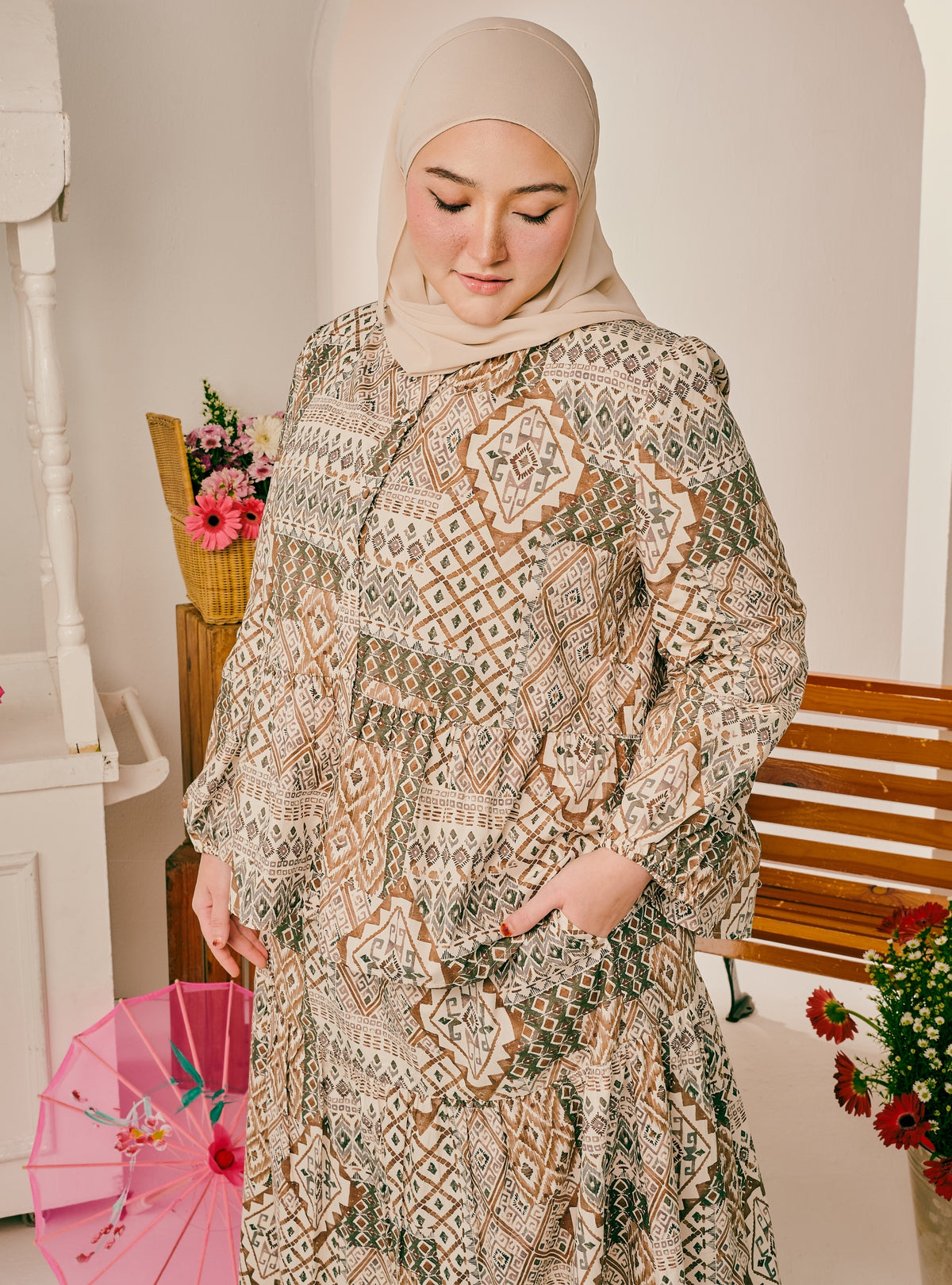 A woman wearing a beige hijab and a long-sleeved, patterned maxi abaya with pockets, standing in a softly lit indoor setting with a wooden bench and pink flowers.