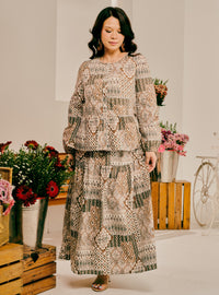A woman modeling a two-piece patchwork maxi dress with a geometric and bohemian pattern in beige, brown, and green tones, set against a styled studio background with flowers and wooden crates.
