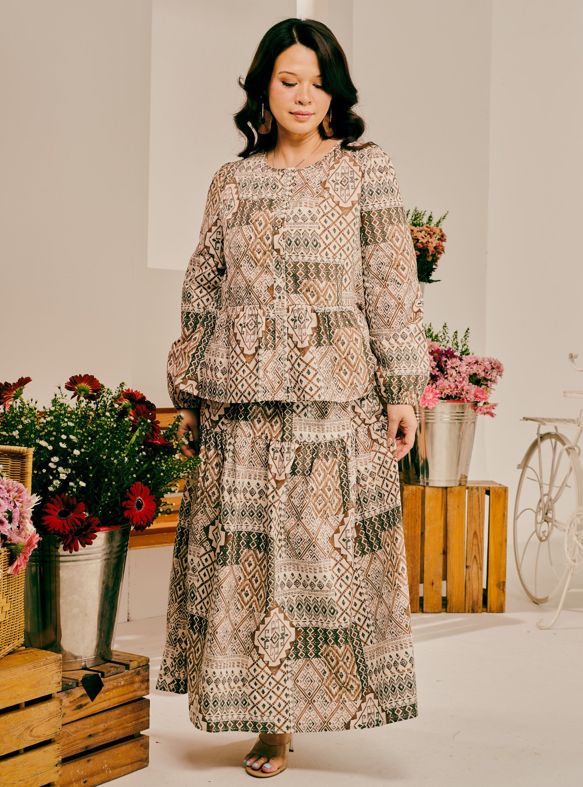 A woman modeling a two-piece patchwork maxi dress with a geometric and bohemian pattern in beige, brown, and green tones, set against a styled studio background with flowers and wooden crates.