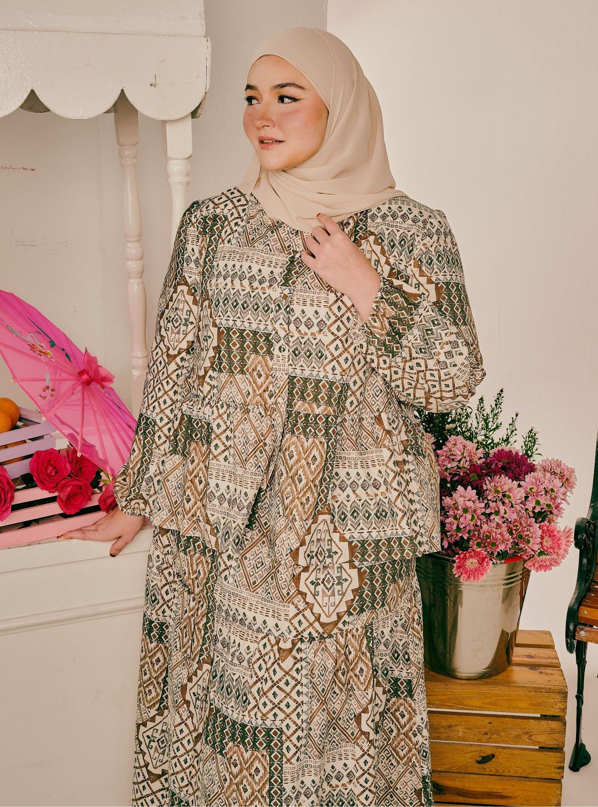 A woman wearing a beige hijab and a long-sleeved, tiered abaya with an intricate geometric patchwork pattern in beige, brown, and green, standing in a decorated indoor setting.