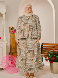 A woman wearing a two-piece modest outfit with a beige, brown, and green geometric pattern, paired with a light beige hijab, standing in a decorated indoor setting with a pink parasol and flowers.