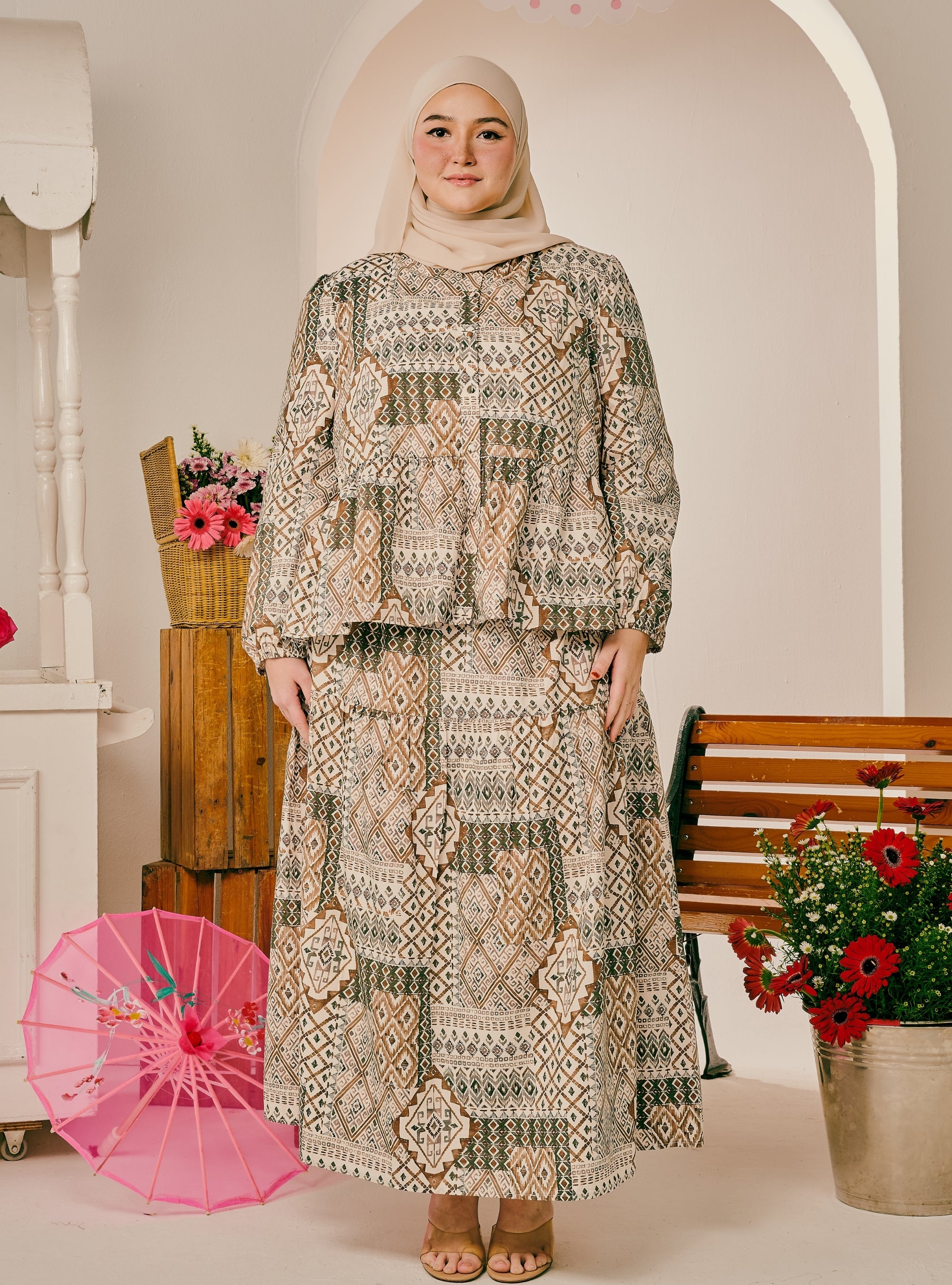 A woman wearing a two-piece modest outfit with a beige, brown, and green geometric pattern, paired with a light beige hijab, standing in a decorated indoor setting with a pink parasol and flowers.
