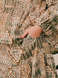 Close-up of a person wearing a tiered long-sleeve dress with a geometric tribal pattern in brown, beige, and green, showing a hand in the side pocket.