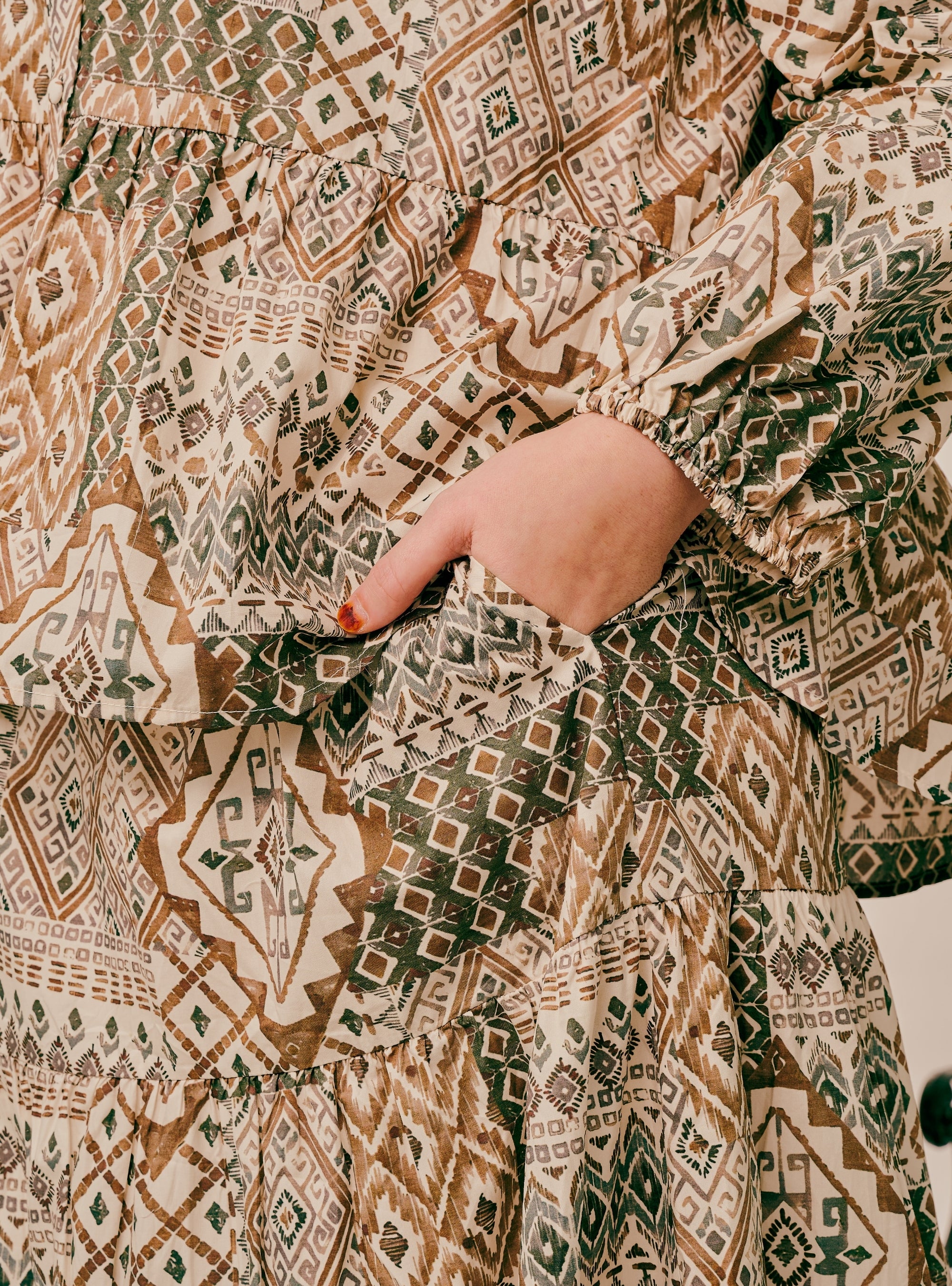 Close-up of a person wearing a tiered long-sleeve dress with a geometric tribal pattern in brown, beige, and green, showing a hand in the side pocket.