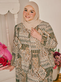 A woman wearing a beige hijab and a long-sleeved, tiered abaya with an intricate geometric and mandala pattern in beige, brown, and olive green. She is standing indoors next to a white wooden chair, holding a pink flower bouquet.