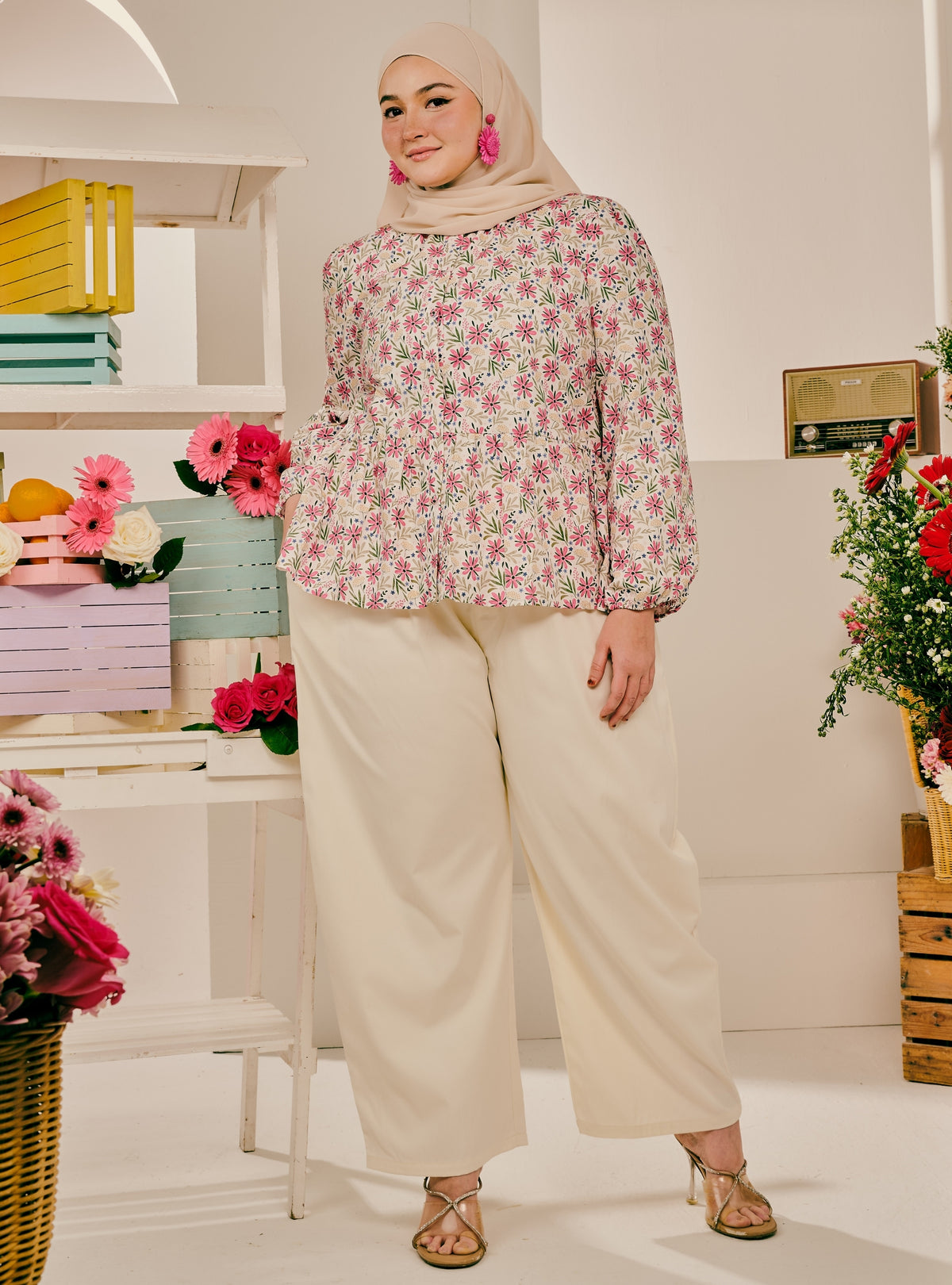 A woman wearing a beige hijab, a pink and green floral long-sleeve blouse, and cream wide-leg trousers, standing in a bright, styled room with shelves and flowers.