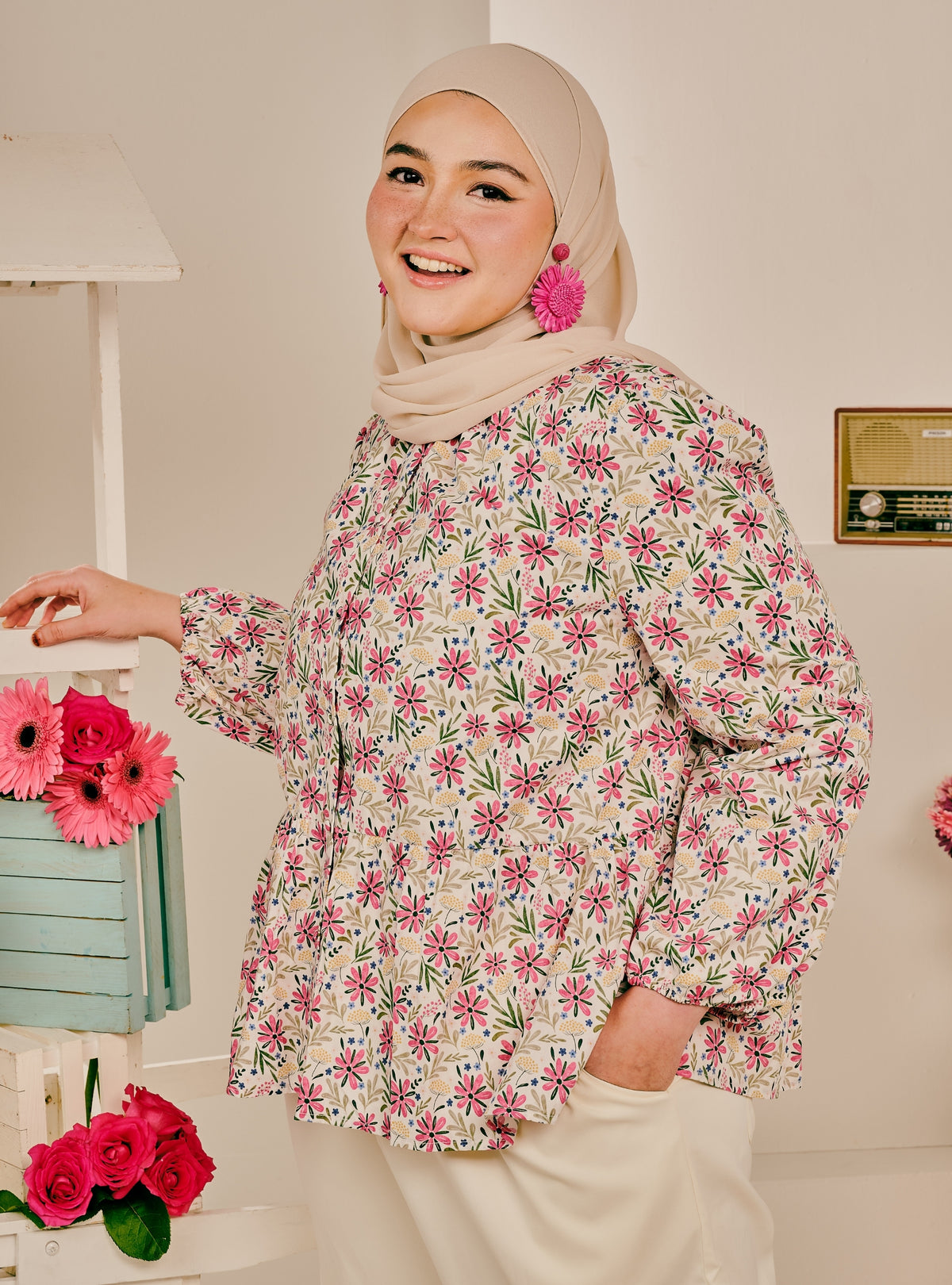 A woman wearing a beige hijab and a long-sleeve floral blouse with cream, pink, and green tones, paired with cream trousers. She is standing indoors next to a decorative shelf with pink flowers.