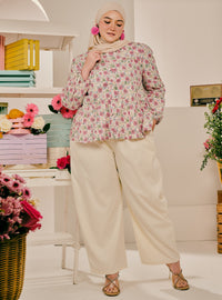 A woman wearing a long-sleeve floral print blouse with beige wide-leg pants, a beige hijab, and gold sandals, standing in a styled indoor room with shelves and flowers.