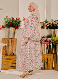 A woman wearing a beige hijab and a long-sleeved, floor-length floral dress with pink and green flowers on a white background, standing in a decorated room with wooden crates and flower arrangements.