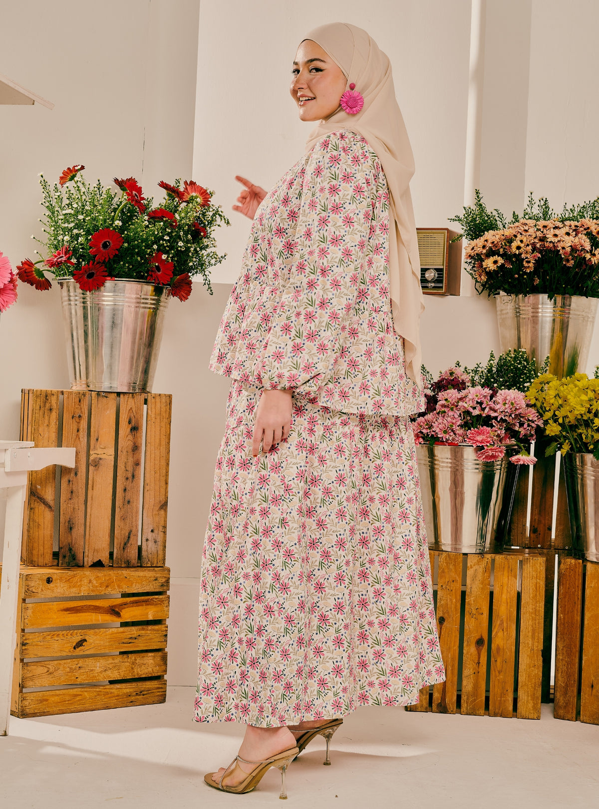 A woman wearing a beige hijab and a long-sleeved, floor-length floral dress with pink and green flowers on a white background, standing in a decorated room with wooden crates and flower arrangements.