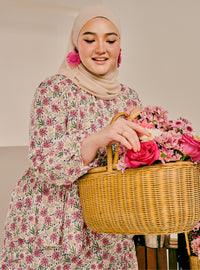 A woman wearing a beige hijab and a long-sleeve floral dress with pink and green flowers, holding a large wicker basket filled with pink roses and flowers.