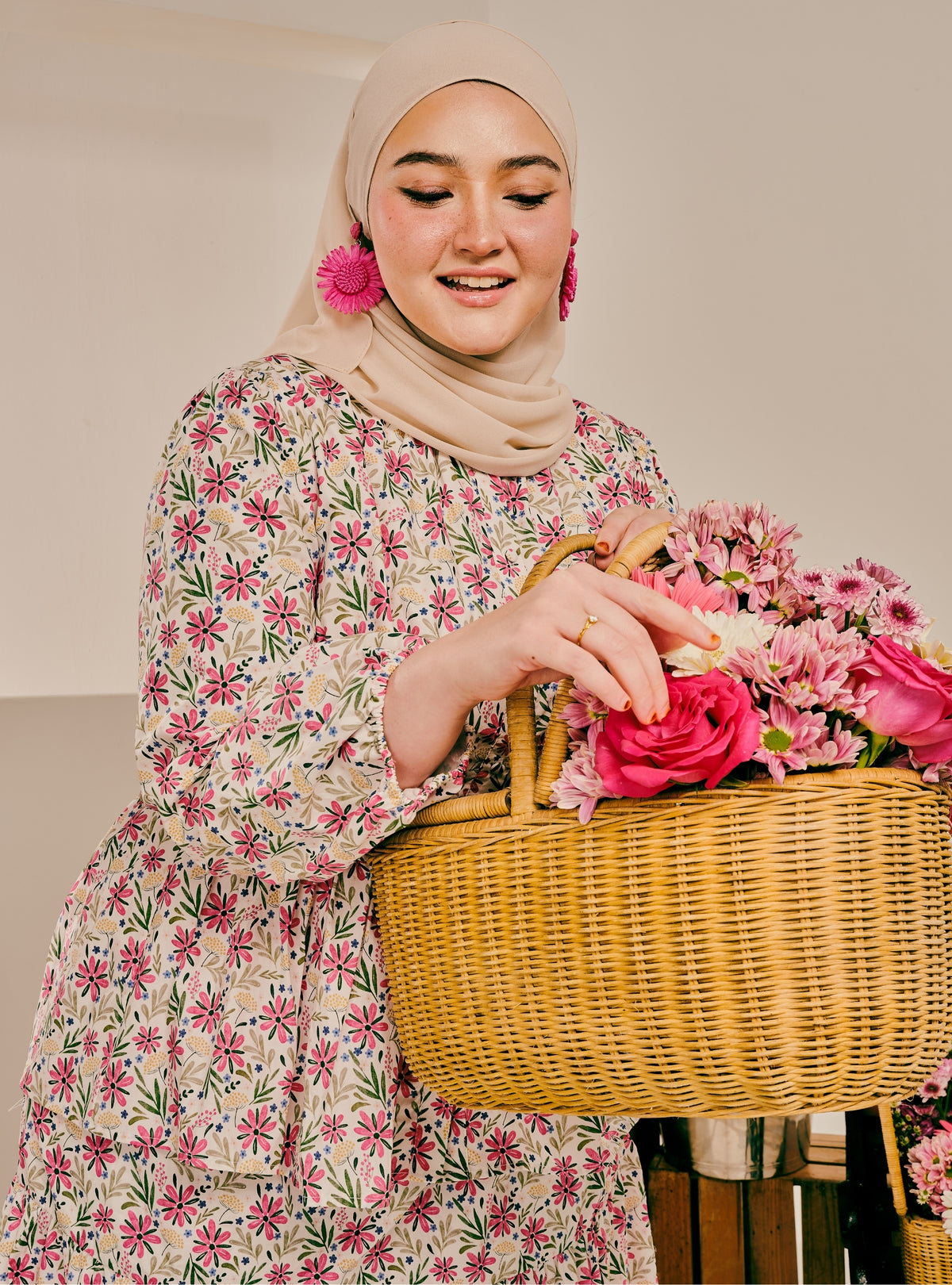 A woman wearing a beige hijab and a long-sleeve floral dress with pink and green flowers, holding a large wicker basket filled with pink roses and flowers.