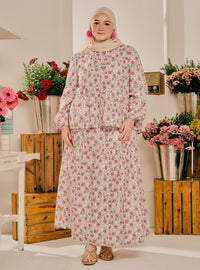 A woman wearing a long-sleeved, high-neck maxi dress with a pink and green floral pattern, paired with a beige hijab and flat sandals, standing in a decorated indoor setting with wooden crates and flower arrangements.