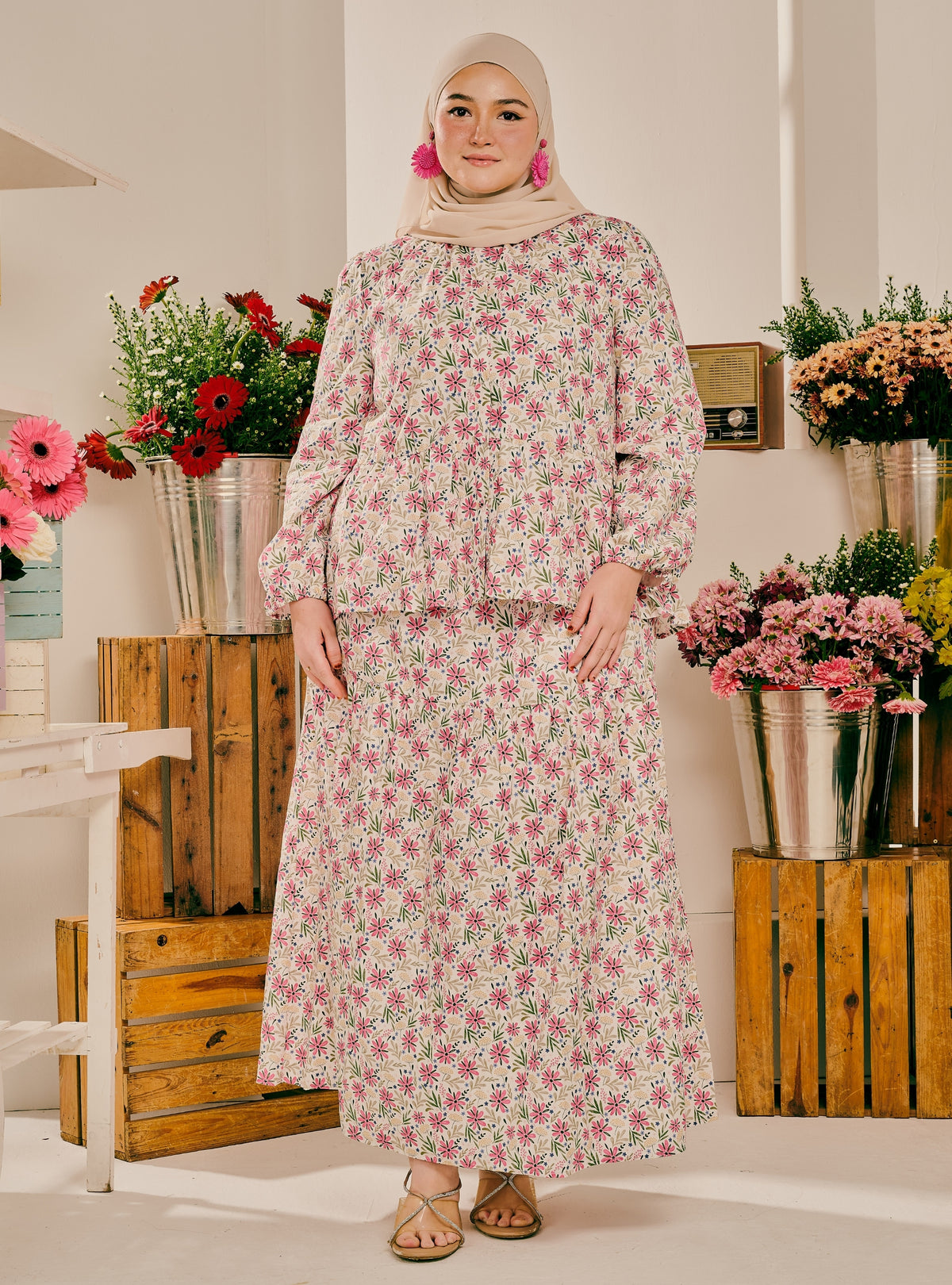 A woman wearing a long-sleeved, high-neck maxi dress with a pink and green floral pattern, paired with a beige hijab and flat sandals, standing in a decorated indoor setting with wooden crates and flower arrangements.