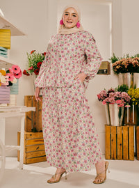 A woman wearing a two-piece floral print modest outfit with a matching hijab, standing in a decorated boutique setting with flowers and wooden crates.