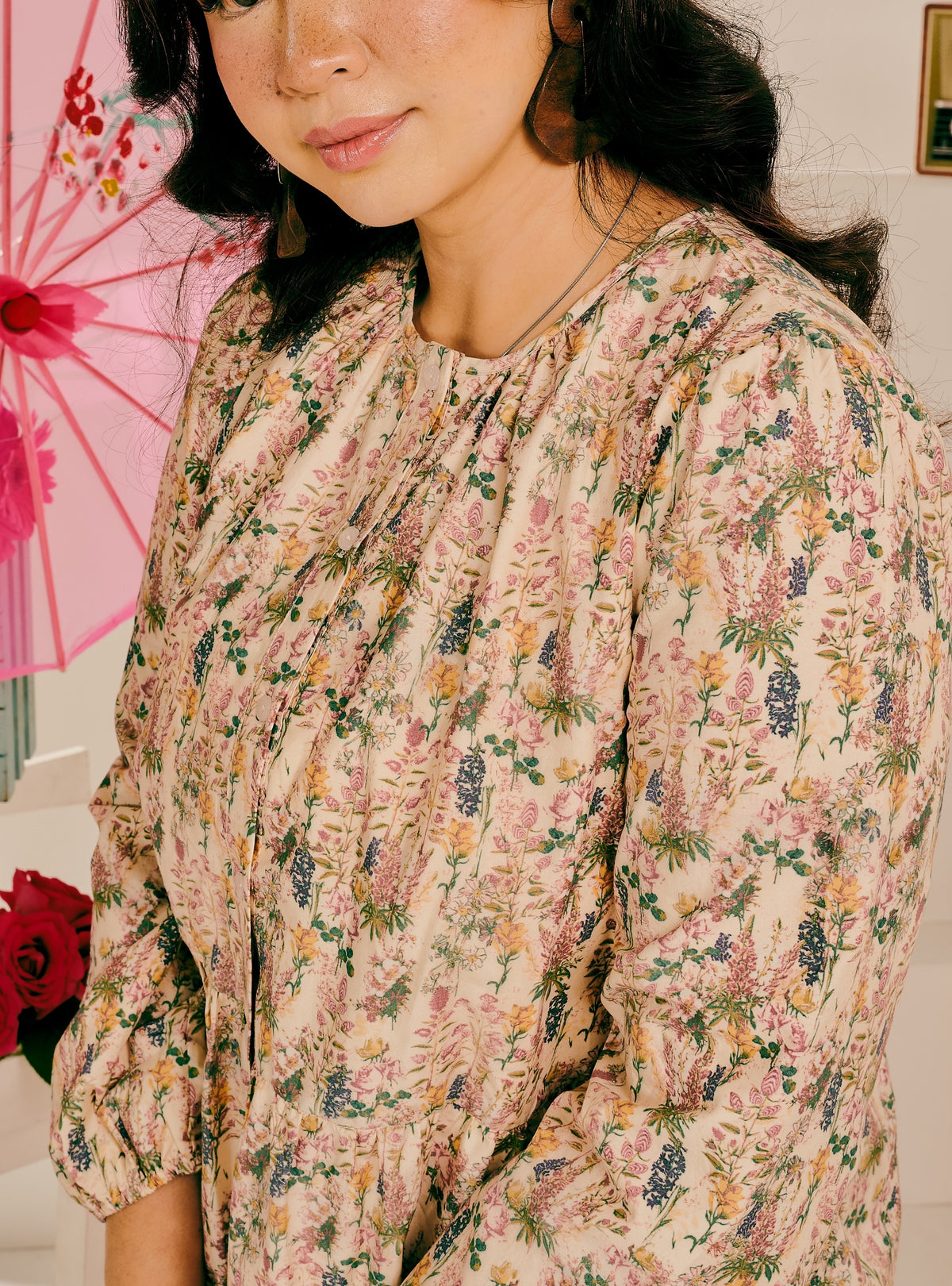 A close-up of a woman wearing a long-sleeve blouse with a dense floral print on a light beige background. The print features pink, purple, yellow, and green botanical motifs.