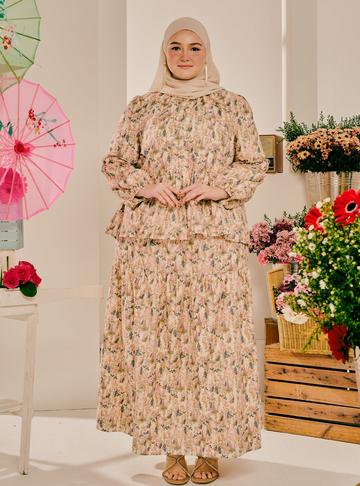 A woman wearing a beige and pink floral print long-sleeve maxi dress with a matching hijab stands in a styled indoor setting with flowers and a pink umbrella in the background.