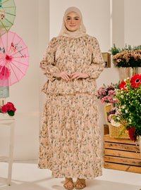 A woman wearing a beige and pink floral print long-sleeve maxi dress with a matching hijab stands in a styled indoor setting with flowers and a pink umbrella in the background.