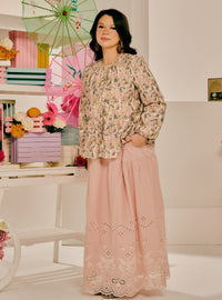 A woman models a two-piece traditional outfit featuring a beige and pink floral printed long-sleeve blouse paired with a matching dusty pink skirt with embroidered eyelet lace at the hem, set against a styled studio background with pastel props.